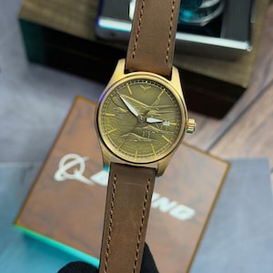 May include: A gold-coloured wristwatch with a brown leather strap. The watch face features a detailed airplane design. The watch has a date display at the 4 o'clock position. The strap is stitched with a classic design.
