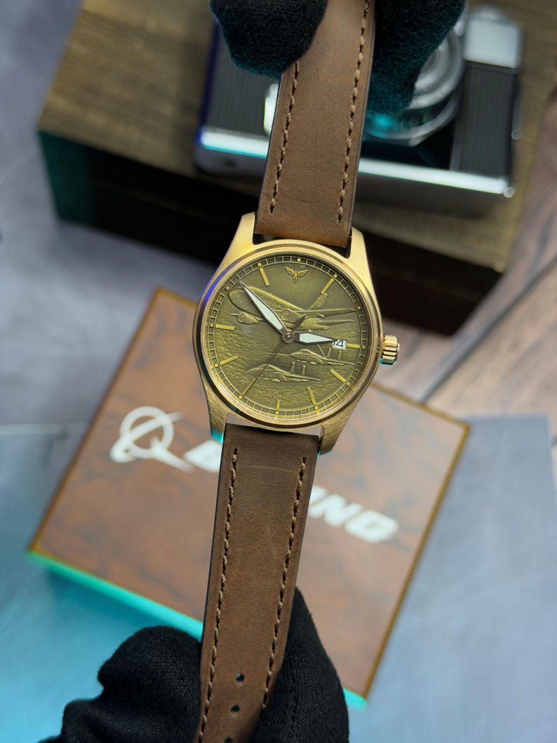May include: A gold-coloured wristwatch with a brown leather strap. The watch face features a detailed airplane design. The watch has a date display at the 4 o'clock position. The strap is stitched with a classic design.