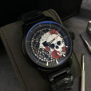 May include: Black wristwatch with a skull and playing card face design. The watch face features a white skull, red roses, and playing cards. The watch has a black strap and a black case.