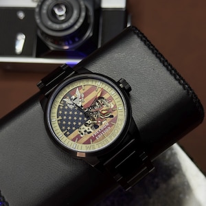 K&ouml;nnte beinhalten: Schwarze Armbanduhr mit einem Zifferblatt mit der amerikanischen Flagge und den Worten "WE THE PEOPLE" und "FREEDOM EDITION". Die Uhr liegt auf einem schwarzen Lederportemonnaie, im Hintergrund eine Vintage-Kamera.