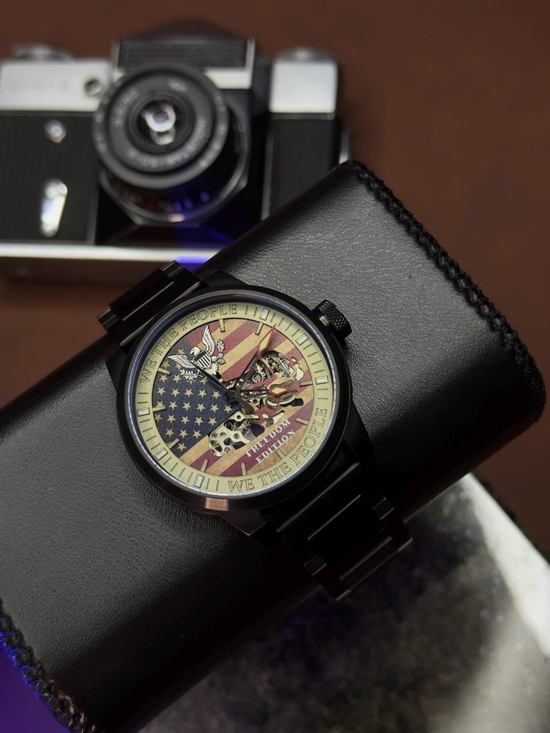 K&ouml;nnte beinhalten: Schwarze Armbanduhr mit einem Zifferblatt mit der amerikanischen Flagge und den Worten "WE THE PEOPLE" und "FREEDOM EDITION". Die Uhr liegt auf einem schwarzen Lederportemonnaie, im Hintergrund eine Vintage-Kamera.