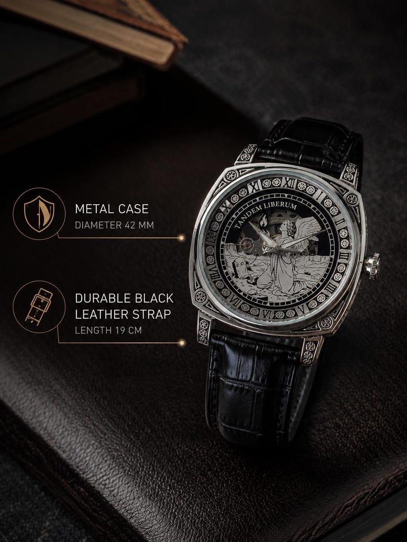 Puede incluir: Un reloj de pulsera plateado con correa de cuero negro y caja de metal de 42 mm. La esfera presenta una ilustraci&oacute;n grabada de un &aacute;ngel, el texto TANDEM LIBERUM y n&uacute;meros romanos. La correa tiene una longitud de 19 cm en total.