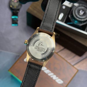 May include: A gold-coloured watch with a black leather strap. The watch face is visible, with the text "SAPPHIRE CRYSTAL AUTO" and "ALL STAINLESS STEEL". The watch is held against a wooden box and a document with the text "BEING".