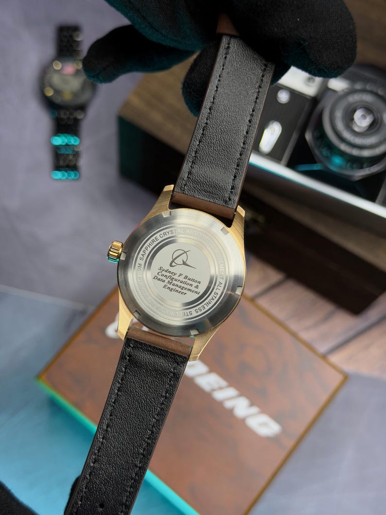 May include: A gold-coloured watch with a black leather strap. The watch face is visible, with the text "SAPPHIRE CRYSTAL AUTO" and "ALL STAINLESS STEEL". The watch is held against a wooden box and a document with the text "BEING".