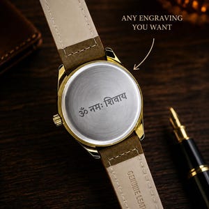 K&ouml;nnte beinhalten: Eine goldfarbene Armbanduhr mit braunem Lederarmband, auf dem "GENUINE LEATHER" und "22" steht. Die R&uuml;ckseite des Uhrengeh&auml;uses ist mit dem Sanskrit-Text "ॐ नमः शिवाय" graviert und enth&auml;lt den Text "ANY ENGRAVING YOU WANT".