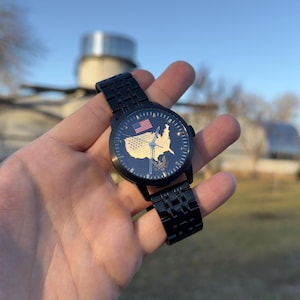 May include: Black wristwatch with a dark blue face featuring a gold-colored map of the United States and a small American flag. The watch has a black metal band and is held in a hand against a blurred outdoor background.
