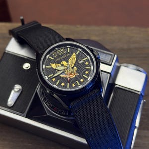 K&ouml;nnte beinhalten: Schwarze Armbanduhr mit strukturiertem schwarzem Armband. Das Zifferblatt zeigt ein goldenes Adleremblem, Sterne und den Text "IN GOD WE TRUST LIMITED EDITION". Die Uhr liegt auf einer Vintage-Kamera.