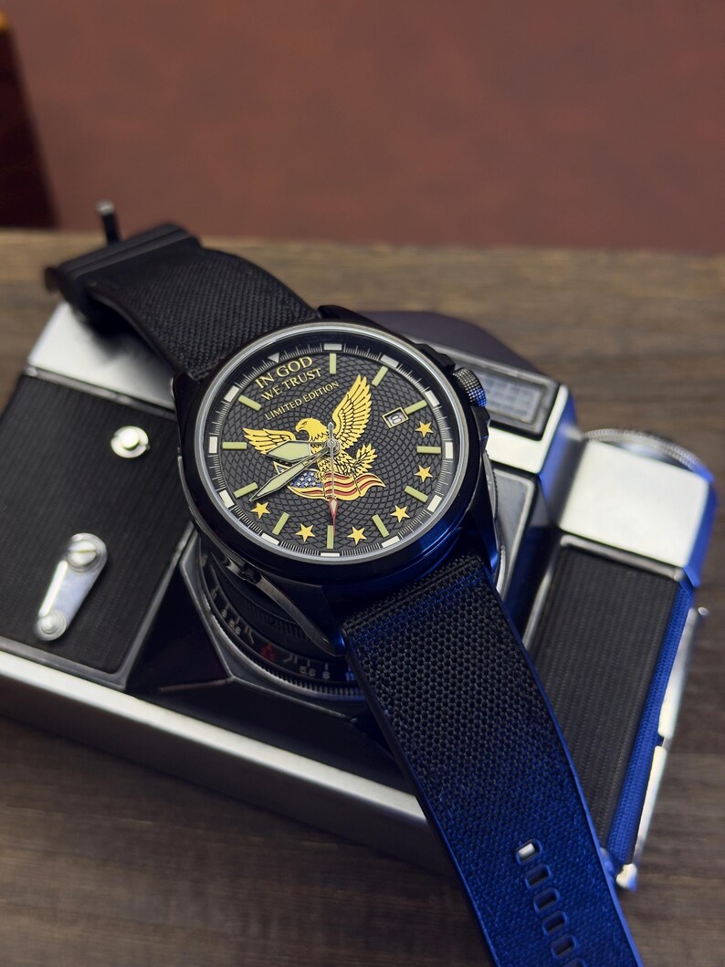 K&ouml;nnte beinhalten: Schwarze Armbanduhr mit strukturiertem schwarzem Armband. Das Zifferblatt zeigt ein goldenes Adleremblem, Sterne und den Text "IN GOD WE TRUST LIMITED EDITION". Die Uhr liegt auf einer Vintage-Kamera.