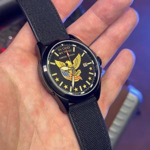 K&ouml;nnte beinhalten: Schwarze Armbanduhr mit schwarzem Armband. Das Zifferblatt zeigt einen goldenen Adler, die amerikanische Flagge und den Text "IN GOD WE TRUST" und "LIMITED EDITION". Die Uhr hat eine Datumsanzeige.