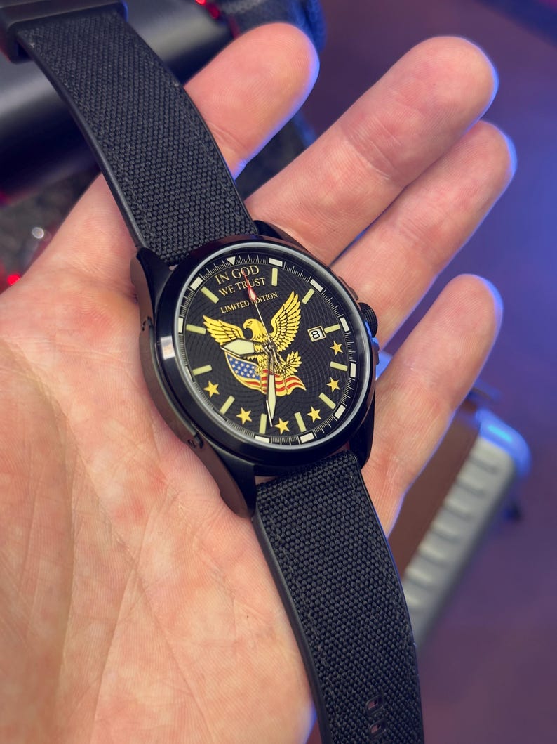 K&ouml;nnte beinhalten: Schwarze Armbanduhr mit schwarzem Armband. Das Zifferblatt zeigt einen goldenen Adler, die amerikanische Flagge und den Text "IN GOD WE TRUST" und "LIMITED EDITION". Die Uhr hat eine Datumsanzeige.