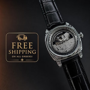 Puede incluir: Un reloj de pulsera plateado con correa de cuero negro con textura de cocodrilo. La esfera muestra una ilustraci&oacute;n detallada de un &aacute;ngel, el texto TANDEM LIBERUM y n&uacute;meros romanos a lo largo del borde del bisel circular.