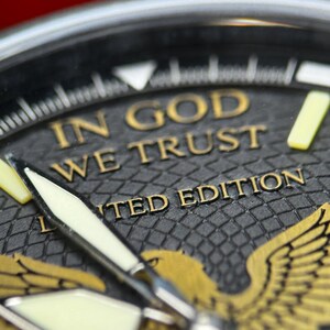 K&ouml;nnte beinhalten: Nahaufnahme eines Zifferblatts mit dem Text "IN GOD WE TRUST" in goldener Schrift. Das Zifferblatt ist schwarz mit einem strukturierten Muster und einem goldenen Adleremblem. Die Worte "LIMITED EDITION" sind ebenfalls sichtbar.