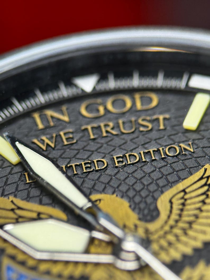 K&ouml;nnte beinhalten: Nahaufnahme eines Zifferblatts mit dem Text "IN GOD WE TRUST" in goldener Schrift. Das Zifferblatt ist schwarz mit einem strukturierten Muster und einem goldenen Adleremblem. Die Worte "LIMITED EDITION" sind ebenfalls sichtbar.