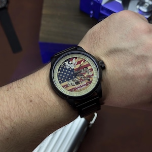 K&ouml;nnte beinhalten: Schwarze Armbanduhr mit patriotischem Design mit der amerikanischen Flagge, einem Adler und dem Text "WE THE PEOPLE". Die Uhr hat ein schwarzes Armband und ein rundes Zifferblatt. Die Uhr ist an einem Handgelenk.