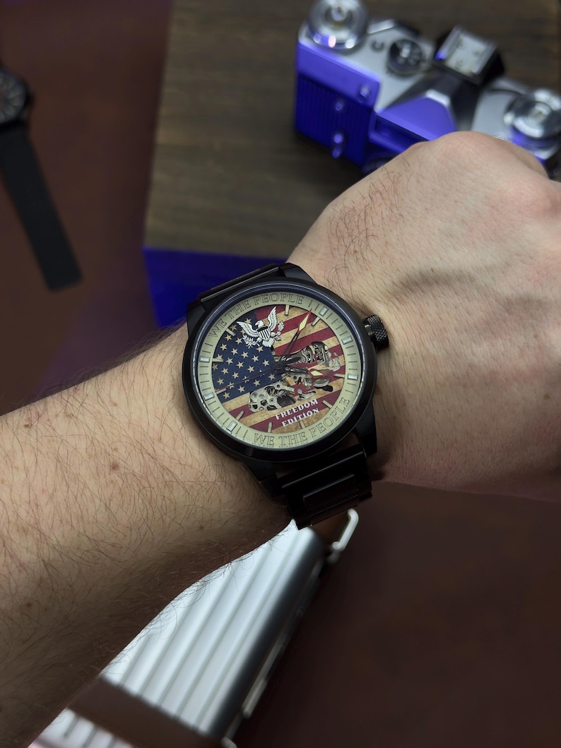 K&ouml;nnte beinhalten: Schwarze Armbanduhr mit patriotischem Design mit der amerikanischen Flagge, einem Adler und dem Text "WE THE PEOPLE". Die Uhr hat ein schwarzes Armband und ein rundes Zifferblatt. Die Uhr ist an einem Handgelenk.