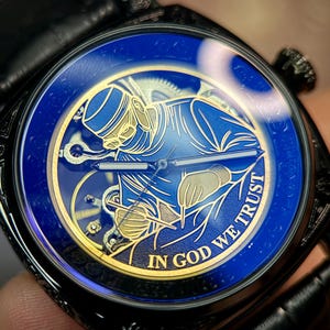 May include: A wristwatch with a black case and leather strap featuring a deep blue dial. The dial displays a gold-toned illustration of a surgeon performing a procedure, with the text IN GOD WE TRUST printed in gold along the bottom edge.
