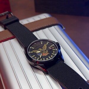 K&ouml;nnte beinhalten: Eine schwarze Armbanduhr mit einem schwarzen Stoffarmband. Das Zifferblatt zeigt einen goldenen Adler, Sterne und den Text "IN GOD WE TRUST LIMITED EDITION". Die Uhr liegt auf einer silbernen und braunen Oberfl&auml;che.