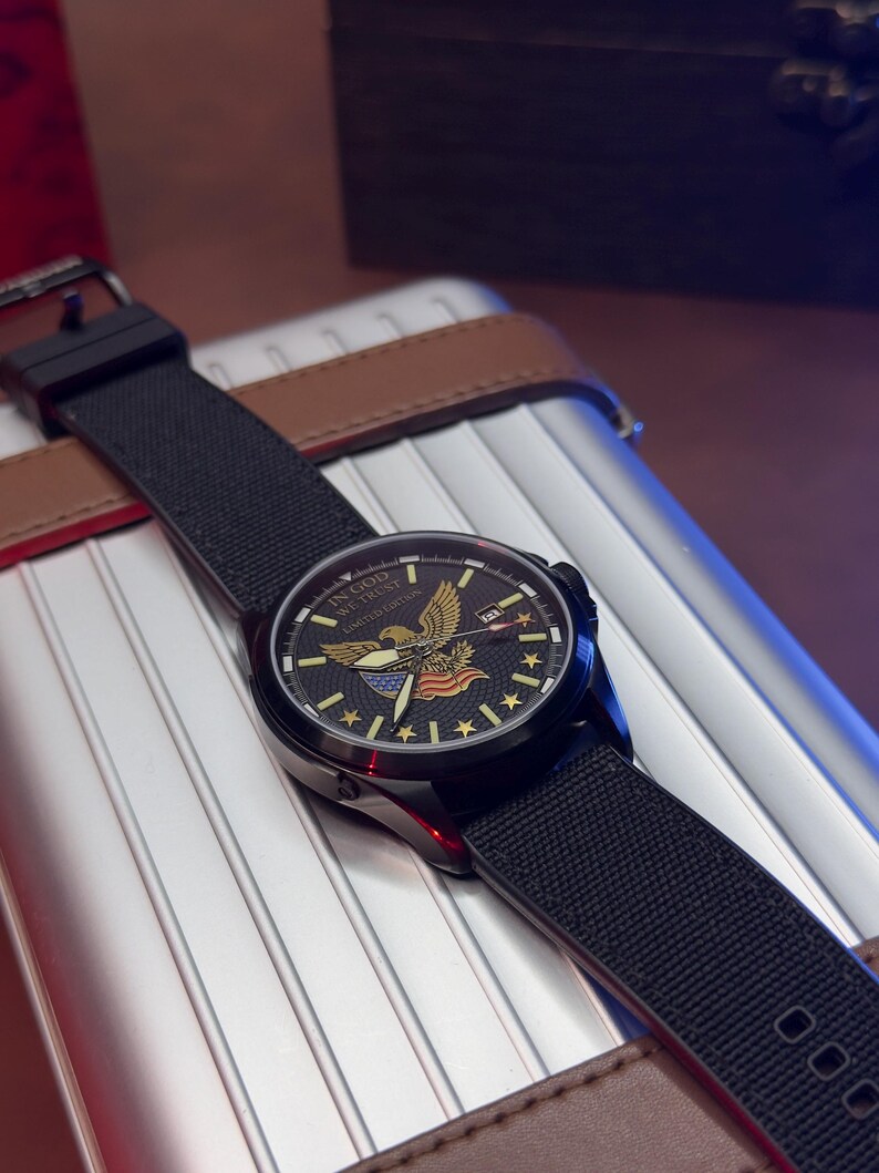 K&ouml;nnte beinhalten: Eine schwarze Armbanduhr mit einem schwarzen Stoffarmband. Das Zifferblatt zeigt einen goldenen Adler, Sterne und den Text "IN GOD WE TRUST LIMITED EDITION". Die Uhr liegt auf einer silbernen und braunen Oberfl&auml;che.