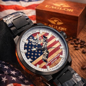 K&ouml;nnte beinhalten: Schwarze Armbanduhr mit einem Zifferblatt mit dem Design der amerikanischen Flagge, einem Adler und dem Text "1776-2026". Die Uhr ist Teil der "250th Anniversary Edition". Die Uhr liegt auf einer Holzoberfl&auml;che mit einer Holzkiste und einer Kaffeetasse im Hintergrund.