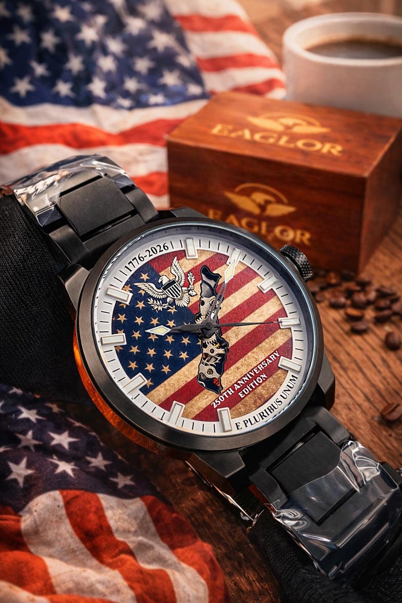 K&ouml;nnte beinhalten: Schwarze Armbanduhr mit einem Zifferblatt mit dem Design der amerikanischen Flagge, einem Adler und dem Text "1776-2026". Die Uhr ist Teil der "250th Anniversary Edition". Die Uhr liegt auf einer Holzoberfl&auml;che mit einer Holzkiste und einer Kaffeetasse im Hintergrund.