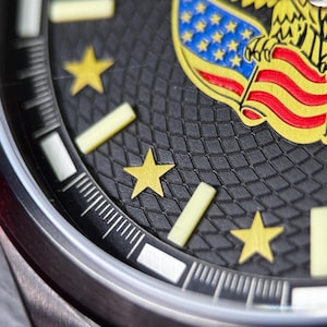 K&ouml;nnte beinhalten: Nahaufnahme eines Armbanduhr-Zifferblatts mit schwarzem strukturiertem Hintergrund. Das Zifferblatt zeigt goldene Sterne, ein gelb-rotes Flaggenmuster und ein silbernes Uhrengeh&auml;use. Die Zeiger der Uhr sind sichtbar.