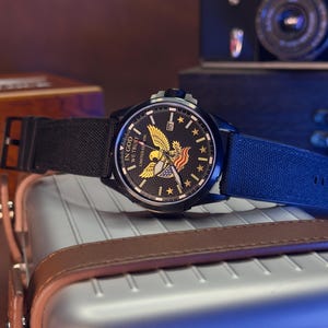 K&ouml;nnte beinhalten: Schwarze Armbanduhr mit Canvas-Armband. Das Zifferblatt zeigt einen goldenen Adler, Sterne und den Text "IN GOD WE TRUST". Die Uhr befindet sich vor einem verschwommenen Hintergrund mit einer Kamera und einem Koffer.