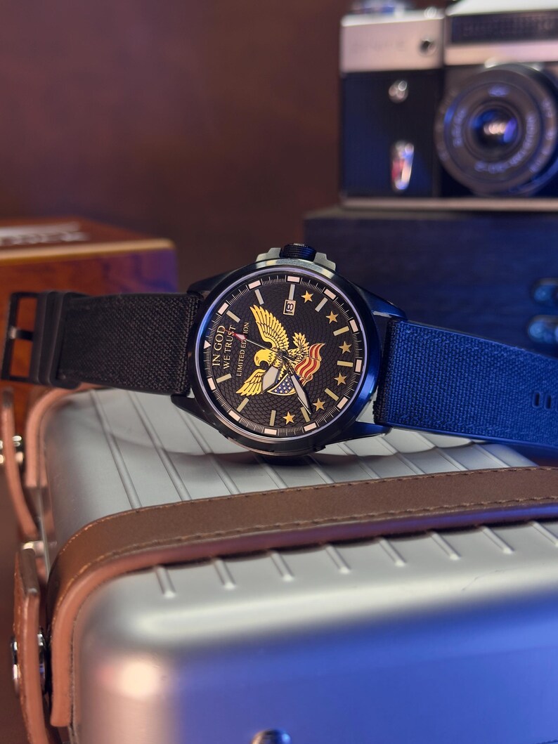 K&ouml;nnte beinhalten: Schwarze Armbanduhr mit Canvas-Armband. Das Zifferblatt zeigt einen goldenen Adler, Sterne und den Text "IN GOD WE TRUST". Die Uhr befindet sich vor einem verschwommenen Hintergrund mit einer Kamera und einem Koffer.