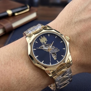 Puede incluir: Reloj de pulsera dorado con una esfera azul oscuro con un diseño de mapa y mecanismos internos intrincados. El reloj tiene una correa dorada y una esfera detallada. El reloj está en una muñeca.