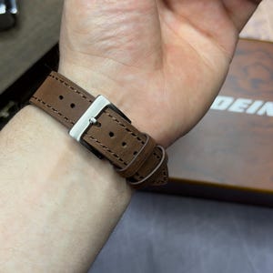 May include: A brown leather watch strap with a silver buckle fastened on a wrist. The strap features visible stitching and a classic design. The watch band is made of a durable material.