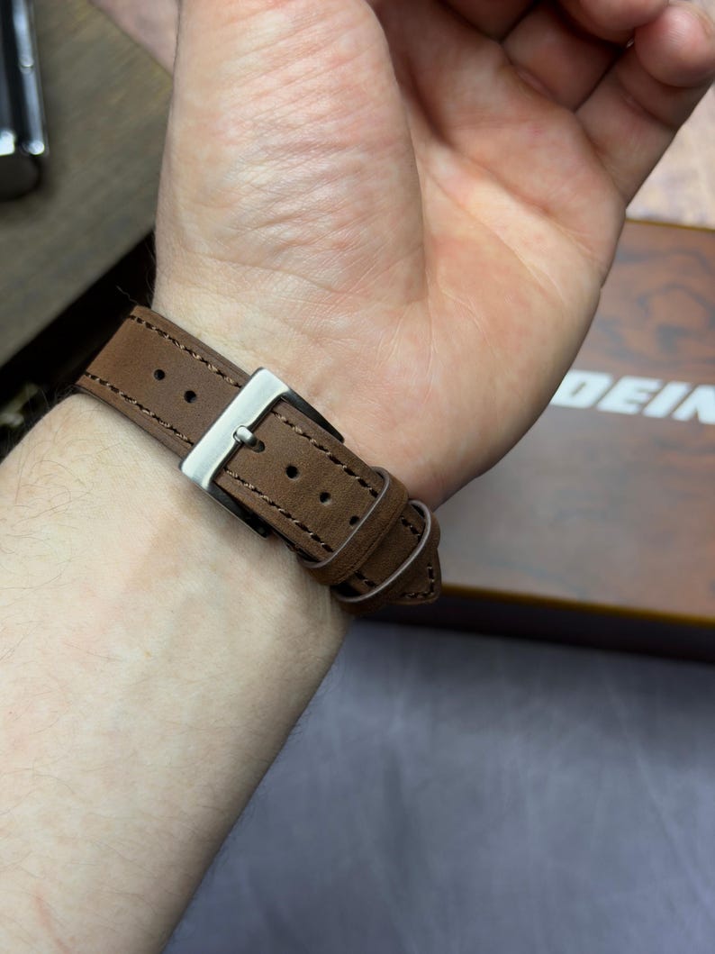 May include: A brown leather watch strap with a silver buckle fastened on a wrist. The strap features visible stitching and a classic design. The watch band is made of a durable material.