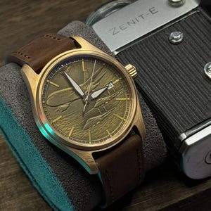May include: A bronze wristwatch with a brown leather strap. The watch face has an engraved airplane design. The watch is displayed on a grey cushion, next to a vintage camera.