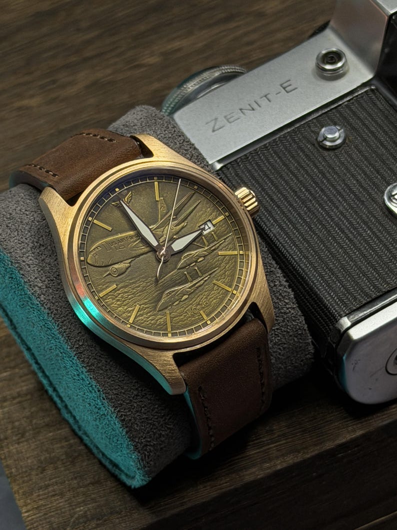 May include: A bronze wristwatch with a brown leather strap. The watch face has an engraved airplane design. The watch is displayed on a grey cushion, next to a vintage camera.