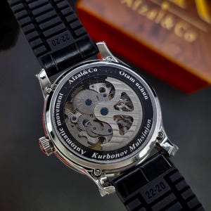 May include: A silver-toned wristwatch with a black rubber strap featuring the text 22-20. The exhibition case back reveals an automatic movement and is engraved with Afzal&Co, Otam uchun, and Kurbonov Muhsinjon in white lettering.