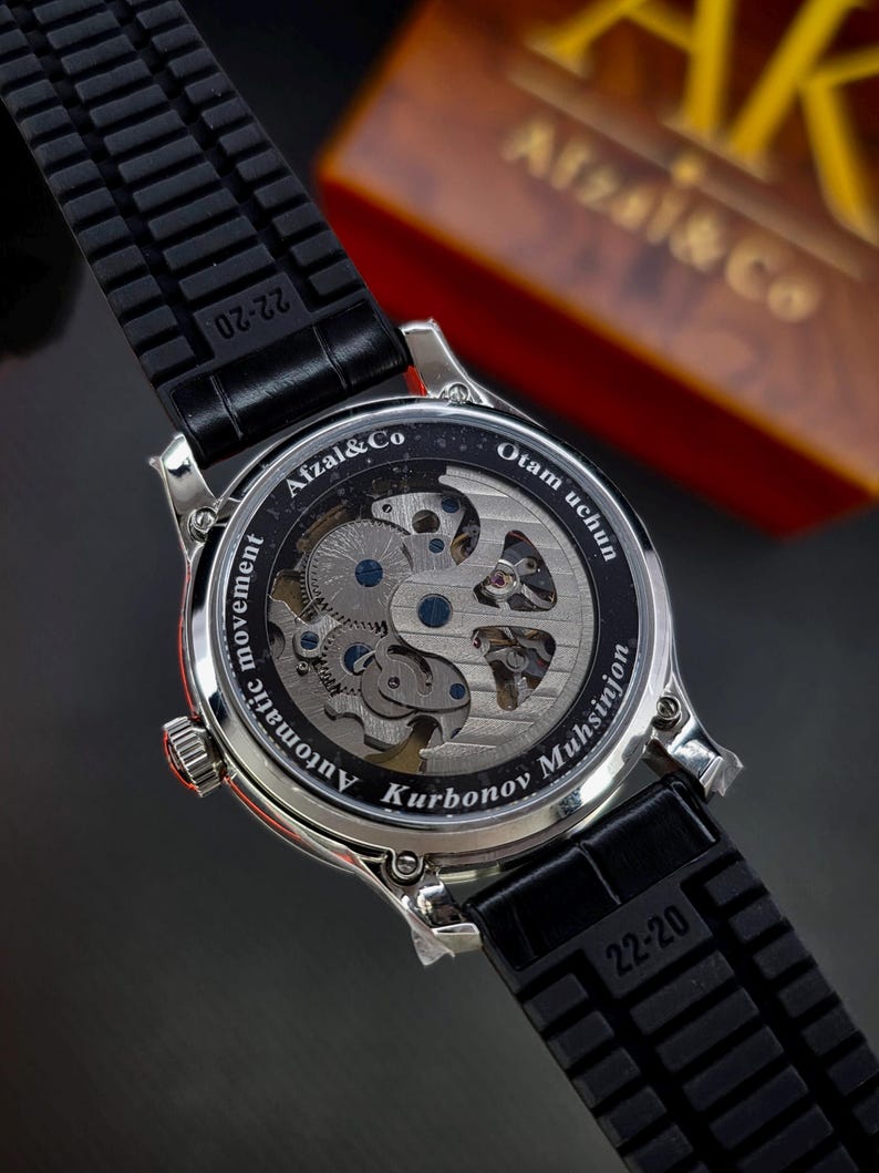 May include: A silver-toned wristwatch with a black rubber strap featuring the text 22-20. The exhibition case back reveals an automatic movement and is engraved with Afzal&Co, Otam uchun, and Kurbonov Muhsinjon in white lettering.