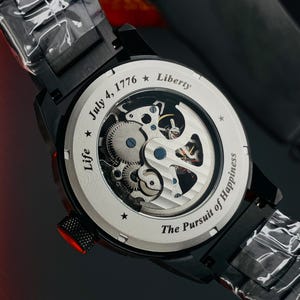K&ouml;nnte beinhalten: Schwarze Armbanduhr mit transparentem R&uuml;cken, die die inneren Mechanismen zeigt. Auf dem Zifferblatt steht "July 4, 1776 * Liberty" und "Life * The Pursuit of Happiness". Die Uhr hat ein schwarzes Armband und eine rote Krone.
