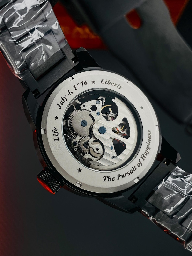 K&ouml;nnte beinhalten: Schwarze Armbanduhr mit transparentem R&uuml;cken, die die inneren Mechanismen zeigt. Auf dem Zifferblatt steht "July 4, 1776 * Liberty" und "Life * The Pursuit of Happiness". Die Uhr hat ein schwarzes Armband und eine rote Krone.