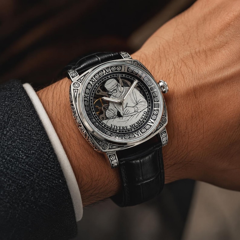 Op de afbeelding: Zilverkleurig polshorloge met een zwarte leren band. De wijzerplaat van het horloge heeft een gedetailleerde gravure van een werkende persoon, omringd door Romeinse cijfers en de woorden "Vivere Memento". Het horloge wordt om de pols gedragen.