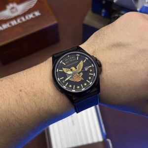 K&ouml;nnte beinhalten: Schwarze Armbanduhr mit einem marineblauen Zifferblatt mit einem Adler, Sternen und dem Text "IN GOD WE TRUST" und "LIMITED EDITION". Die Uhr hat ein schwarzes Armband und wird am Handgelenk getragen. Eine Holzkiste mit dem Text "ARCH CLOCK" befindet sich im Hintergrund.