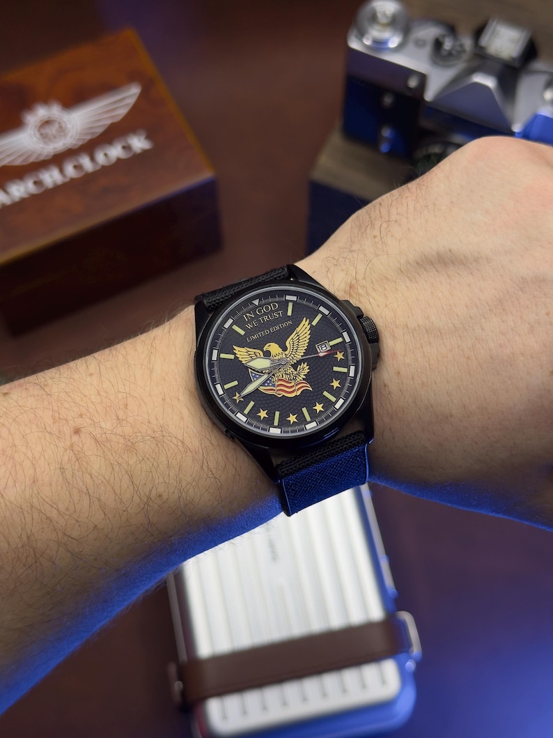 K&ouml;nnte beinhalten: Schwarze Armbanduhr mit einem marineblauen Zifferblatt mit einem Adler, Sternen und dem Text "IN GOD WE TRUST" und "LIMITED EDITION". Die Uhr hat ein schwarzes Armband und wird am Handgelenk getragen. Eine Holzkiste mit dem Text "ARCH CLOCK" befindet sich im Hintergrund.