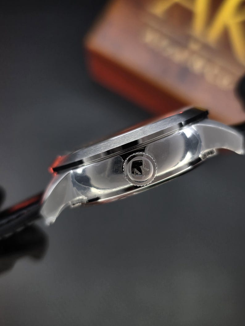 May include: A side view of a silver-toned wristwatch case featuring a polished metallic finish and a textured crown. The crown is engraved with the letters AK. The watch case has a brushed top surface and a smooth, curved side profile.