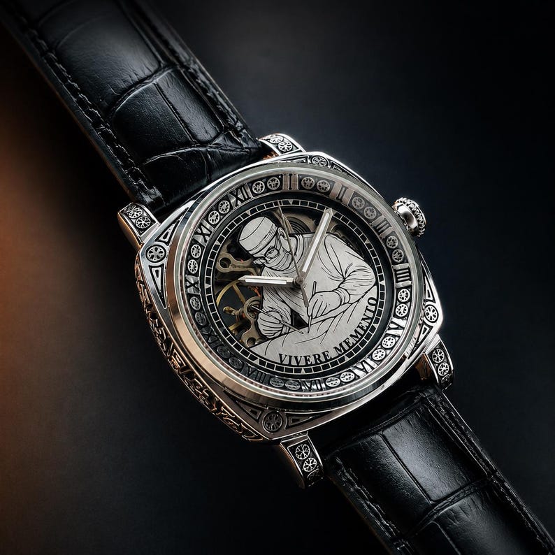 Op de afbeelding: Een zilverkleurig polshorloge met een zwarte leren band. De wijzerplaat heeft een gegraveerde illustratie van een werkende persoon, met de woorden "VIVERE MEMENTO" rond de rand. Het horloge heeft Romeinse cijfers.