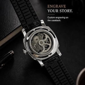 Op de afbeelding: Close-up van een zilverkleurig horloge met een zwarte rubberen band. De achterkant is transparant en toont het ingewikkelde binnenwerk. De tekst "ENGRAVE YOUR STORY" en "Custom engraving on the caseback" zijn zichtbaar. De band is 22 mm breed.