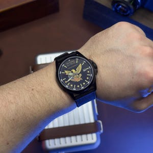 K&ouml;nnte beinhalten: Schwarze Armbanduhr mit dunkelblauem Zifferblatt mit einem Adleremblem und dem Text "IN GOD WE TRUST". Die Uhr hat ein schwarzes Armband und eine Datumsanzeige. Die Uhr ist an einem Handgelenk.