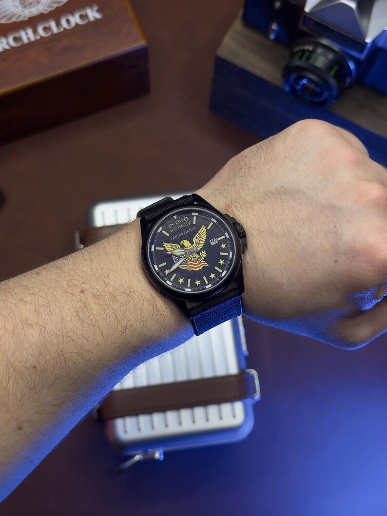 K&ouml;nnte beinhalten: Schwarze Armbanduhr mit dunkelblauem Zifferblatt mit einem Adleremblem und dem Text "IN GOD WE TRUST". Die Uhr hat ein schwarzes Armband und eine Datumsanzeige. Die Uhr ist an einem Handgelenk.