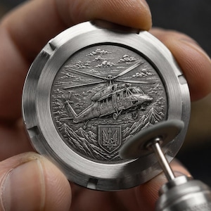 May include: A circular silver-toned metal medallion featuring a detailed relief engraving of a helicopter flying over a mountainous landscape with a trident emblem below. A small rotary tool with a polishing disc is positioned on the edge.