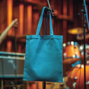 May include: A solid teal tote bag with long handles hangs from a microphone stand. The bag is made of a sturdy fabric and is empty. The background is a blurred stage setting with musical instruments.