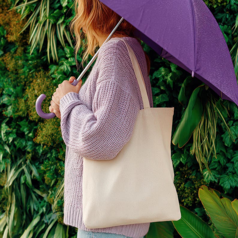May include: A person holding a purple umbrella and a cream-colored tote bag. The person is wearing a light purple sweater. The background is a wall of green plants.