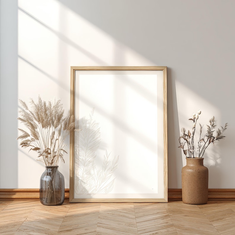 May include: A large, empty framed print leans against a white wall, with a wooden floor. Two vases with dried plants flank the frame. The frame is light wood, and the vases are glass and ceramic. Sunlight casts shadows on the wall.