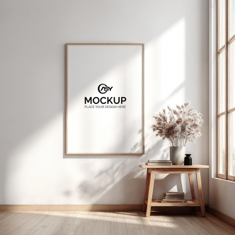 May include: A framed mockup with the text "MOCKUP PLACE YOUR DESIGN HERE" on a white wall. A small wooden table holds a vase of dried flowers, books, and a black decorative bottle. Natural light streams in from a nearby window.
