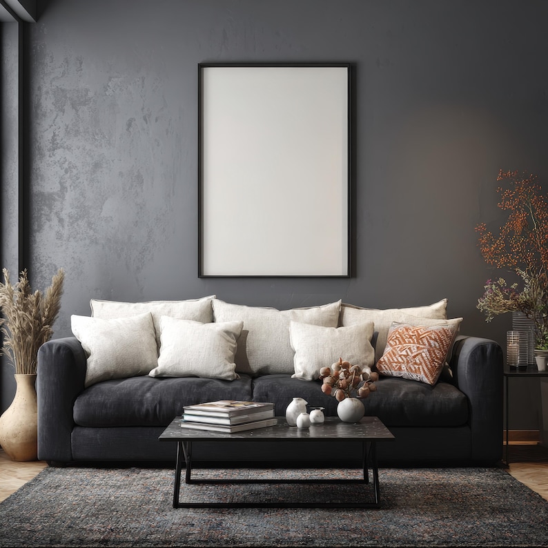 May include: A modern living room with a dark gray sofa, adorned with cream-colored pillows and a patterned orange cushion. A black framed blank canvas hangs above the sofa. A dark gray rug and coffee table complete the scene.